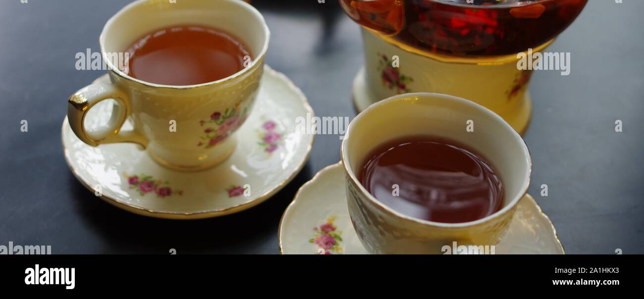 Tè nero con goji (chinese wolfberries) e gelsomino nella teiera in vetro e porcellana tazze con motivo floreale piattini su tavola nera Foto Stock