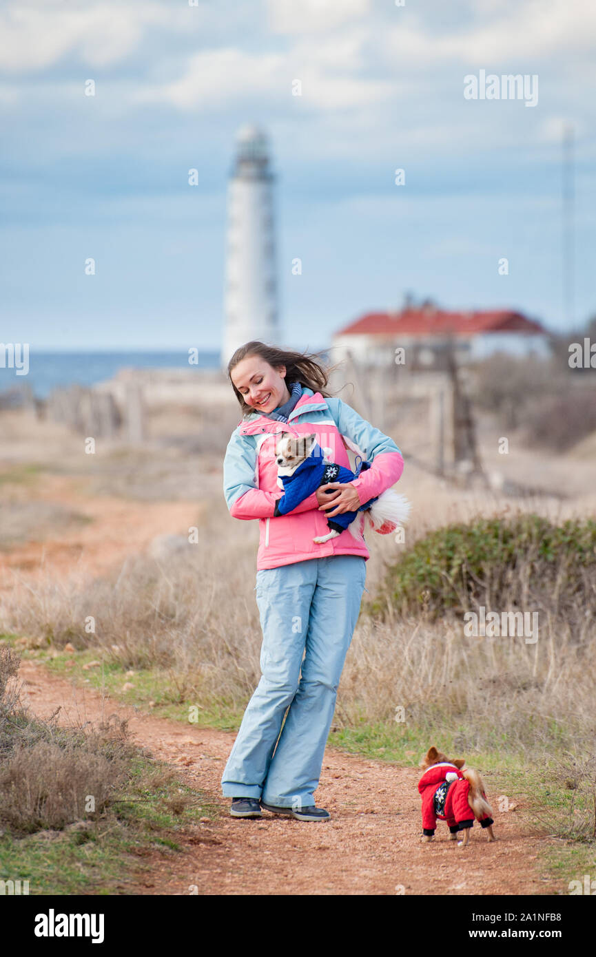 Felice sano attivo deliziosa giovane donna tenendo poco chihuahua cane in abiti animale vicino alla costa dell'oceano con il faro Foto Stock