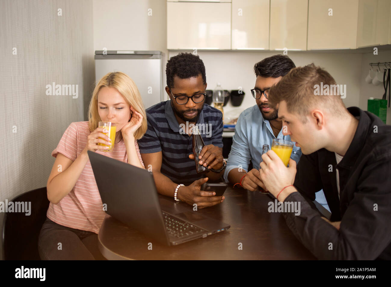 Gruppo di diversi studenti seduti a tavola in cucina, bevendo succo, utilizzando il telefono cellulare e il computer portatile guardando film insieme. Amicizia, svago, TEC Foto Stock