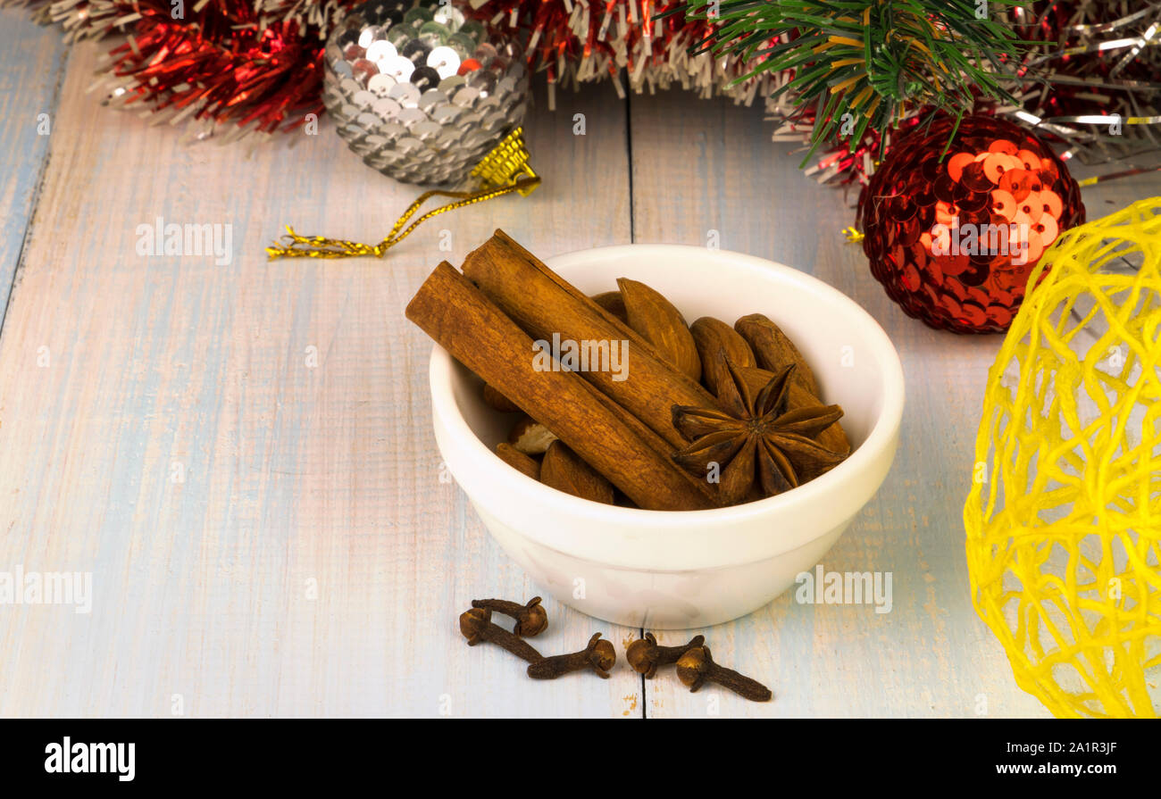 Ciotola bianca con bastoncini di cannella, anice stellato e mandorle e chiodi di garofano su un tavolo di legno, rosso tinsel, finto albero di Natale ramo, giallo snowball di vimini Foto Stock