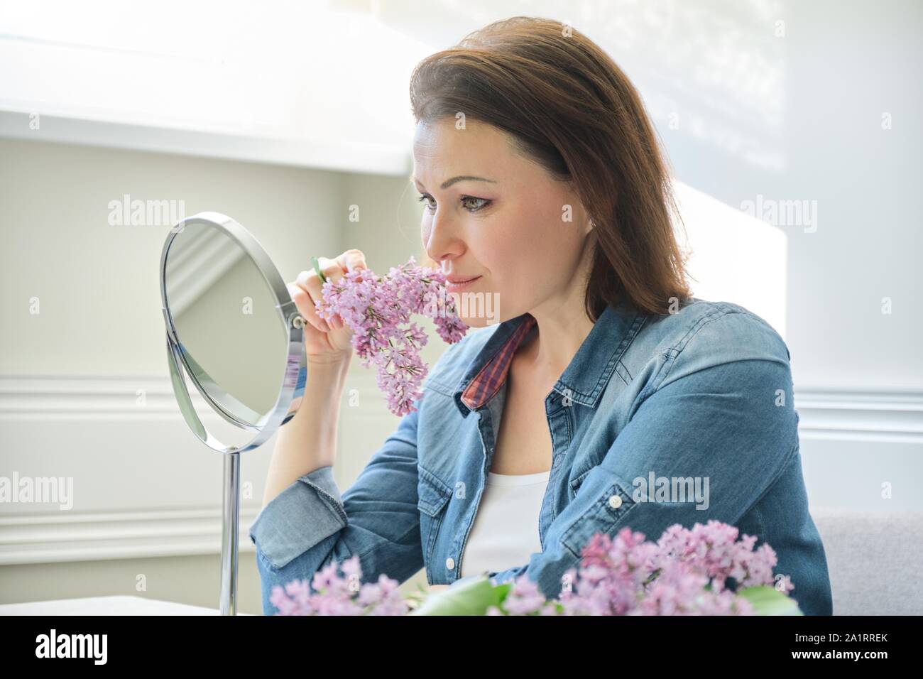Ritratto di donna di mezza età con specchio per il make-up, bella femmina 40 anni seduto a casa a tavola con fiori lilla Foto Stock