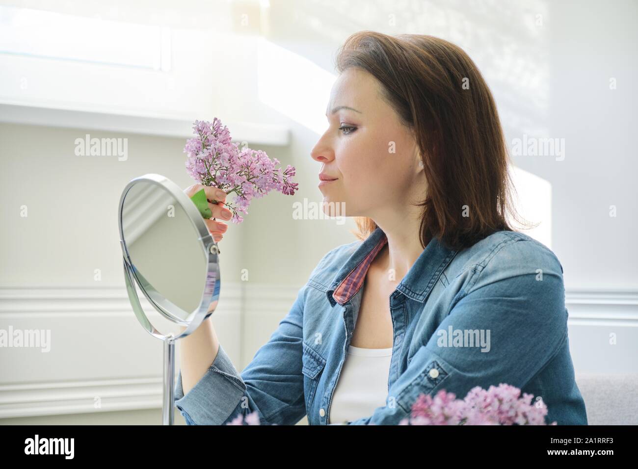 Ritratto di donna di mezza età con specchio per il make-up, bella femmina 40 anni seduto a casa a tavola con fiori lilla Foto Stock