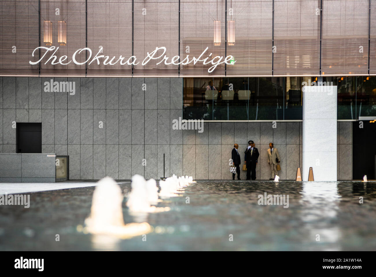 La scritta "The Okura Prestige" brilla sopra l'ingresso del The Okura Tokyo, nel quartiere di Minato, Giappone, con tre portieri in livrea in piedi sul bordo di una vasca d'acqua con fontane illuminate davanti. Foto Stock