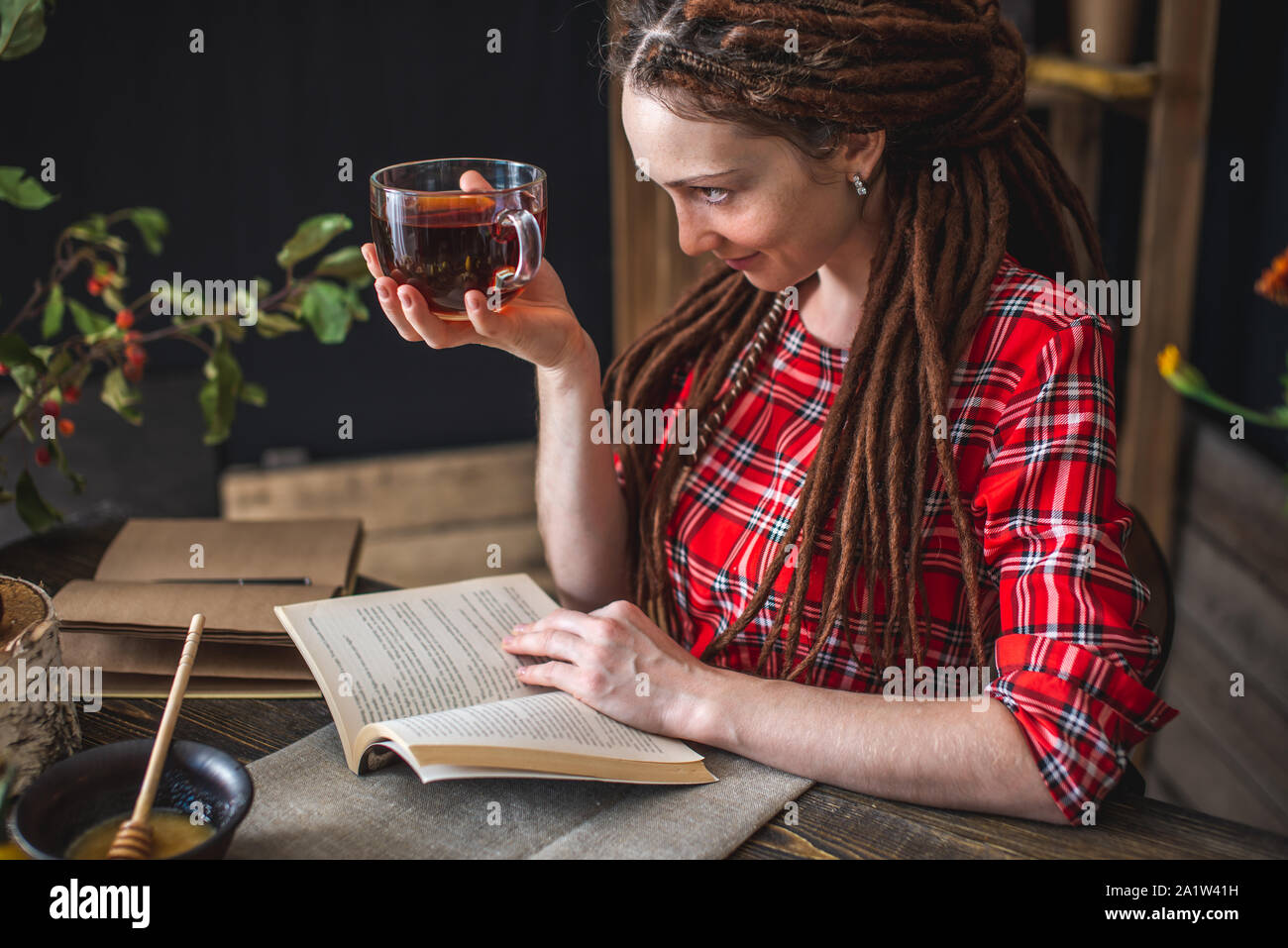 Bella donna la lettura di un libro romantico in un accogliente atmosfera caldo godendo l'autunno dell'umore. Tavolo rustico con fiori di colore giallo e una pila di libri Foto Stock