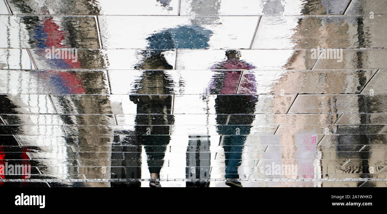 Sfocate silhouette di riflessione sul bagnato via della città di due persone turistiche camminare sotto un ombrellone e tirando in viaggio valigie nella piovosa giornata d'autunno Foto Stock