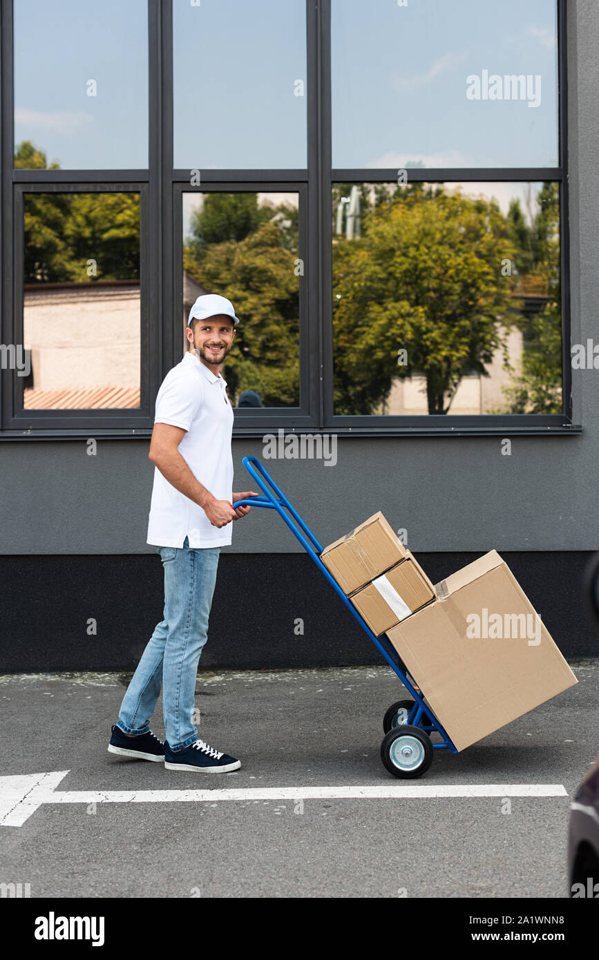 Felice di consegna barbuto uomo in piedi con consegna carrello vicino all'edificio Foto Stock