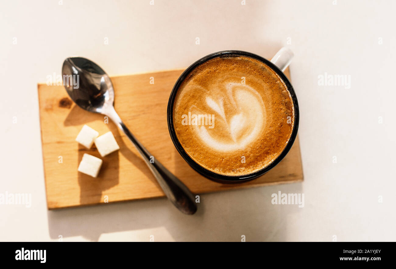 Tazza di caffè latte con schiuma di cui sopra, un cucchiaio di zucchero e su sfondo bianco. Giornata di sole. Foto Stock