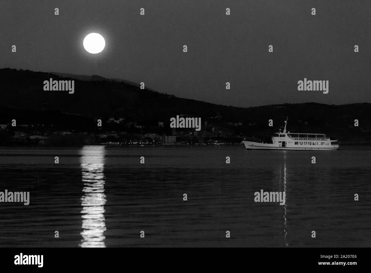 Full Moon Rising su alcune colline del lago Trasimeno, rispecchiando perfettamente in acqua con un traghetto nelle vicinanze Foto Stock