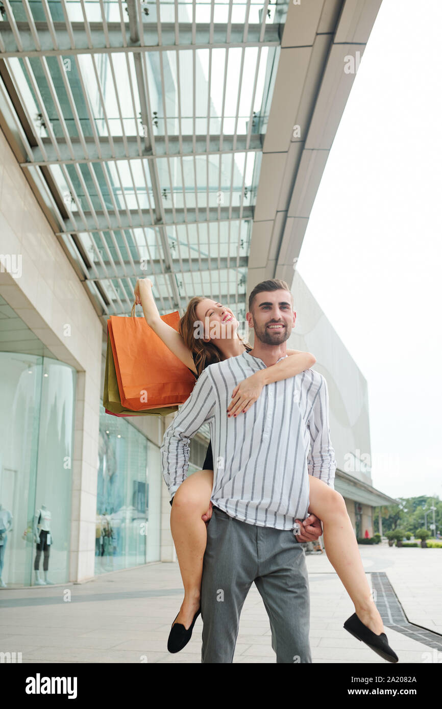 Cherful bel giovane dando piggyback ride alla ragazza dopo lo shopping nel centro commerciale togeter Foto Stock
