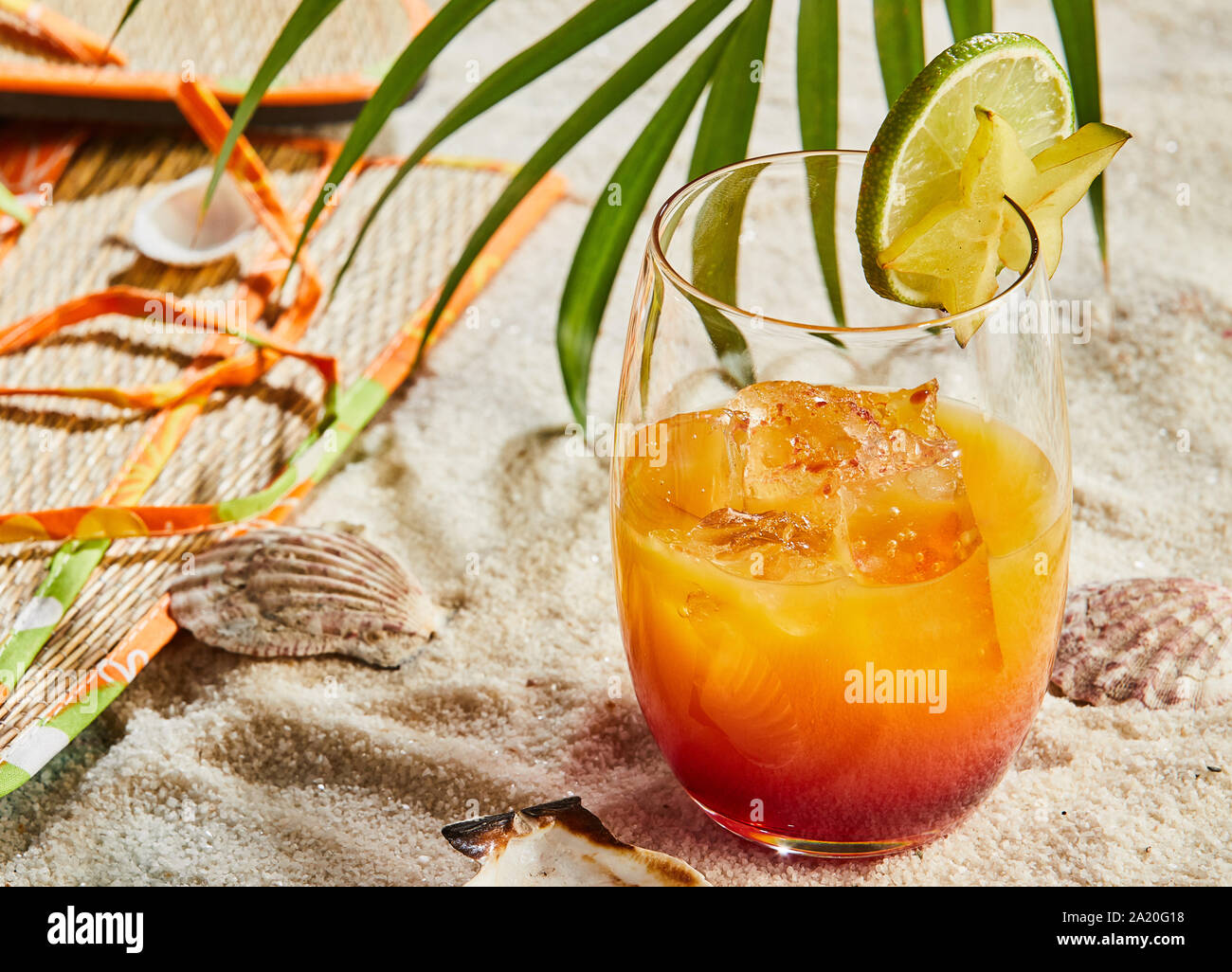 Bicchiere di Orange e rum blend cocktail con ghiaccio guarnita con calce su una sabbiosa spiaggia tropicale sotto una palma Foto Stock