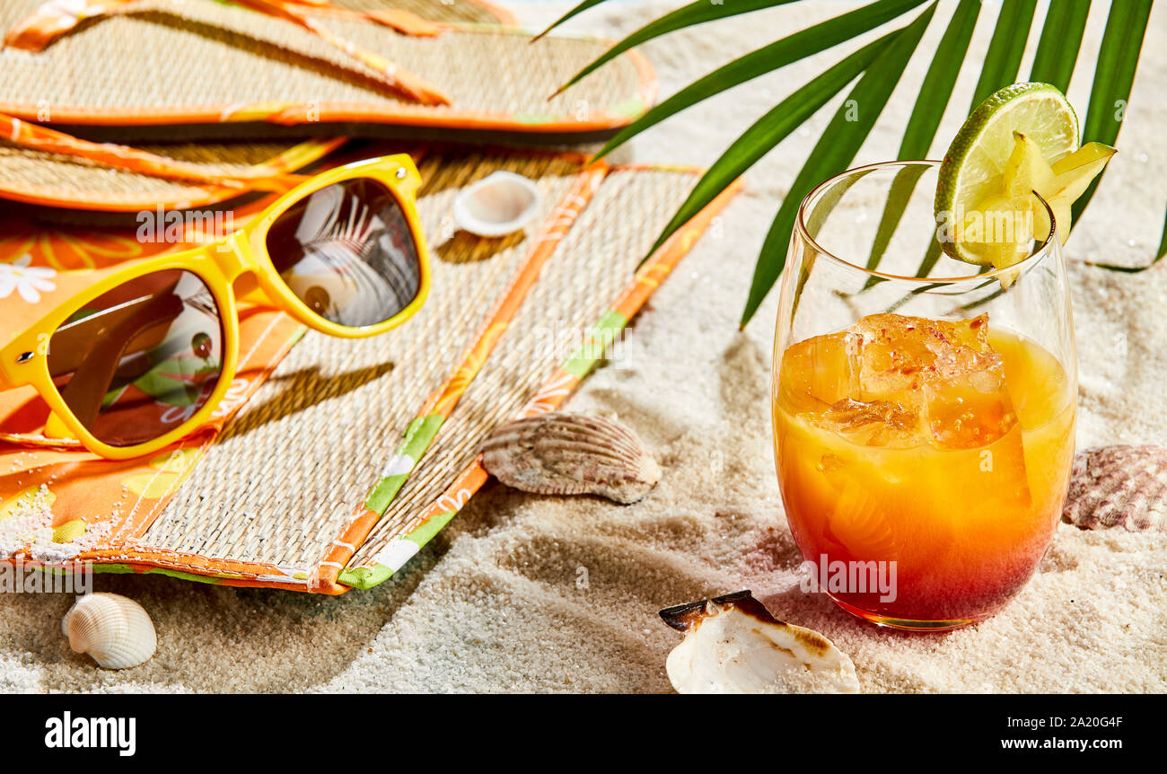 Rum cocktail arancione su una spiaggia tropicale in piedi sulla sabbia bianca in estate il sole a fianco di una borsa da spiaggia, occhiali da sole e sandali Foto Stock