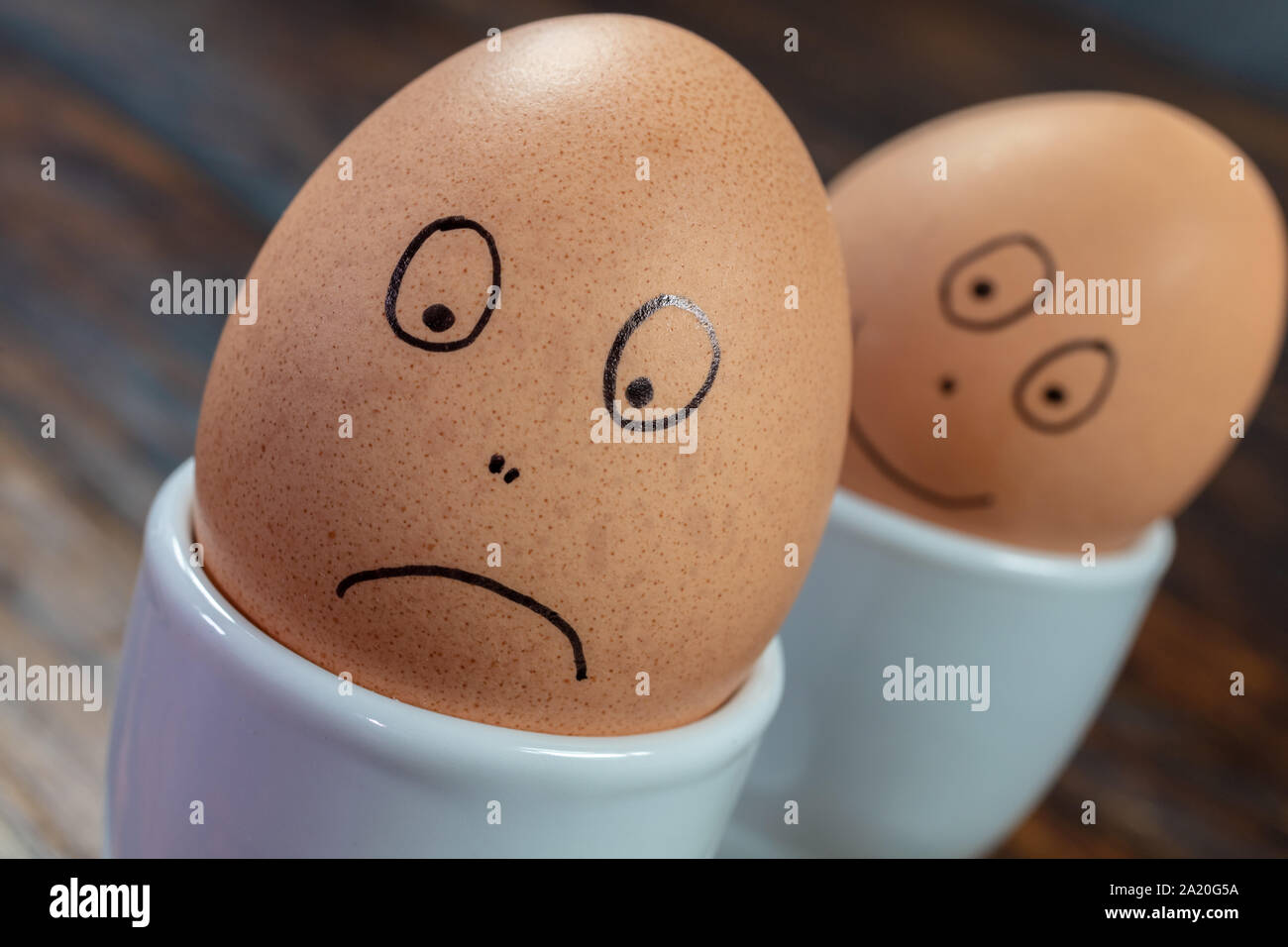 Emozioni concetto due uova sode in bianco d'uovo con coppe disegnate felici e tristi si affaccia su di un tavolo di legno Foto Stock
