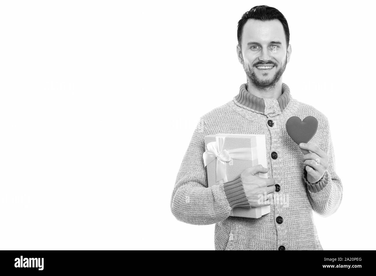 Studio shot di felice giovane uomo sorridente tenendo confezione regalo e cuore rosso pronto per il giorno di San Valentino Foto Stock