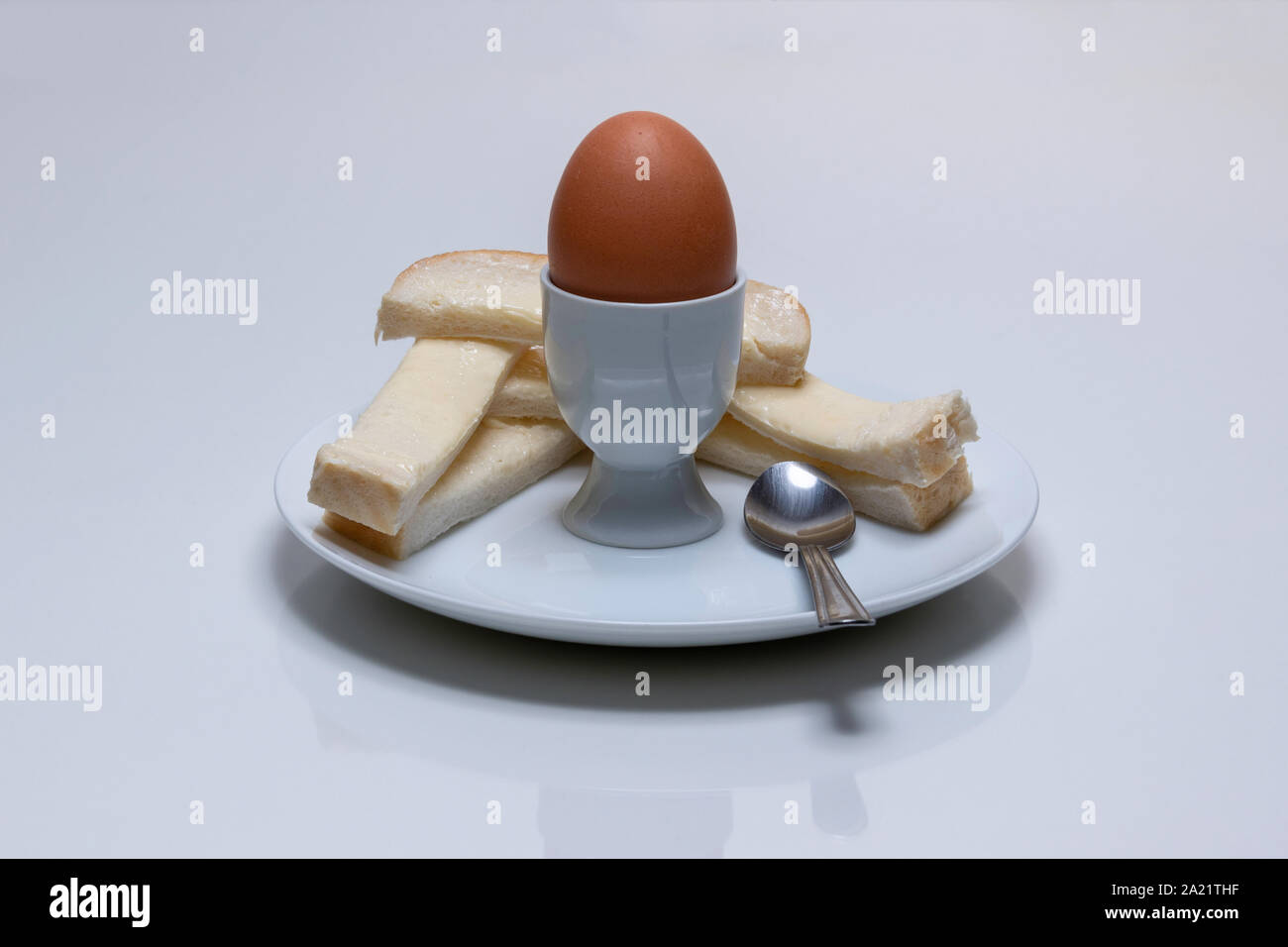 Pollo lesso di uova, in un uovo cup con pane imburrato, soldati e un cucchiaino su una piastra bianca Foto Stock