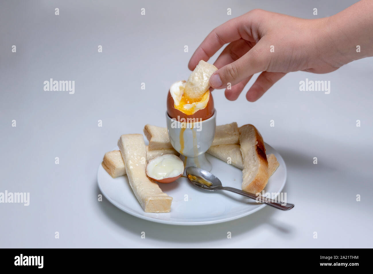 Soft boiled pollo uovo, in un uovo cup con pane imburrato, soldati e un cucchiaio da tè in appoggio su una piastra. Mano intingere il pane soldato nell'uovo Foto Stock