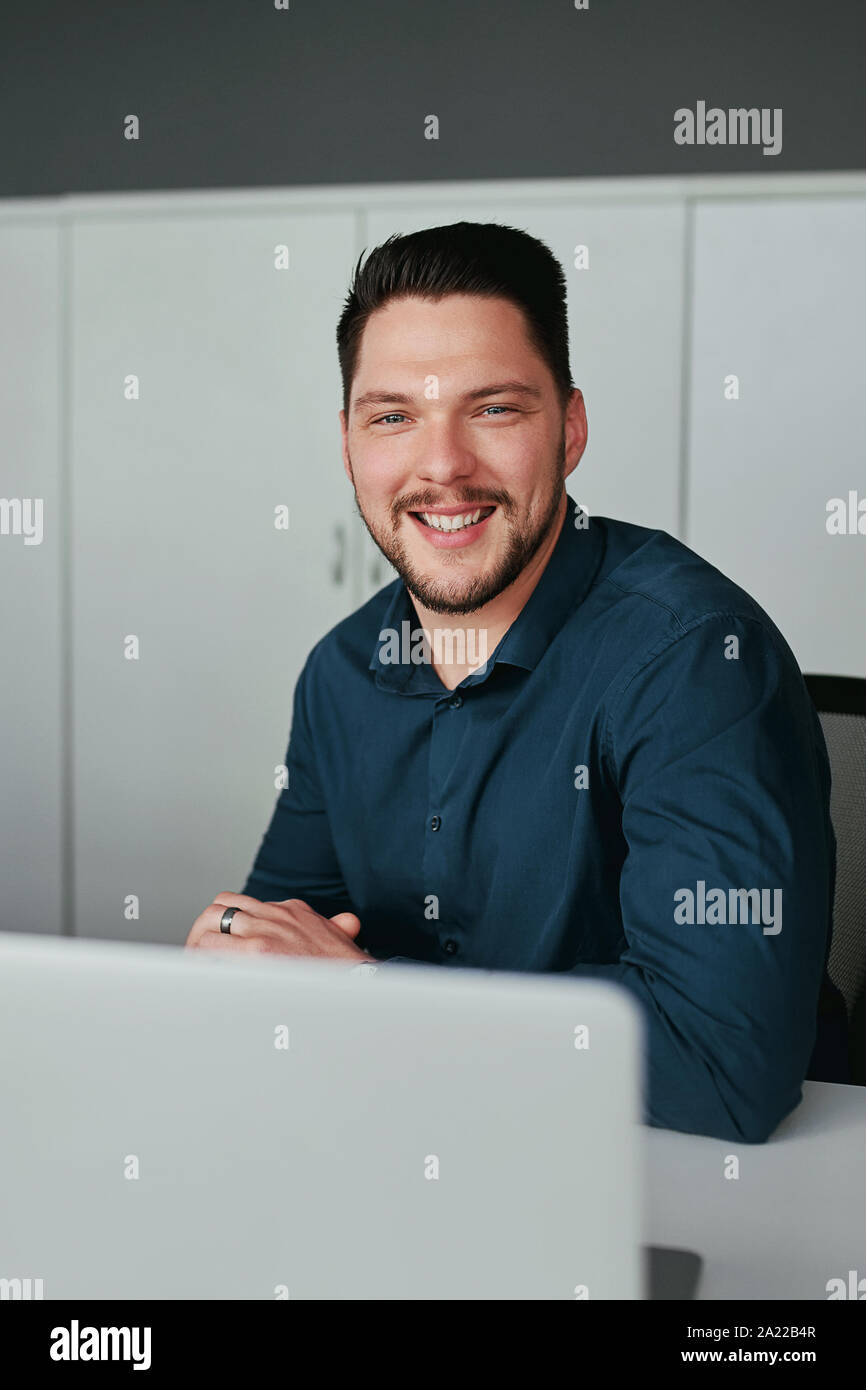 Ritratto di un ben vestito giovane imprenditore sorridente seduta tranquillamente con il computer portatile guardando la fotocamera alla scrivania in ufficio Foto Stock