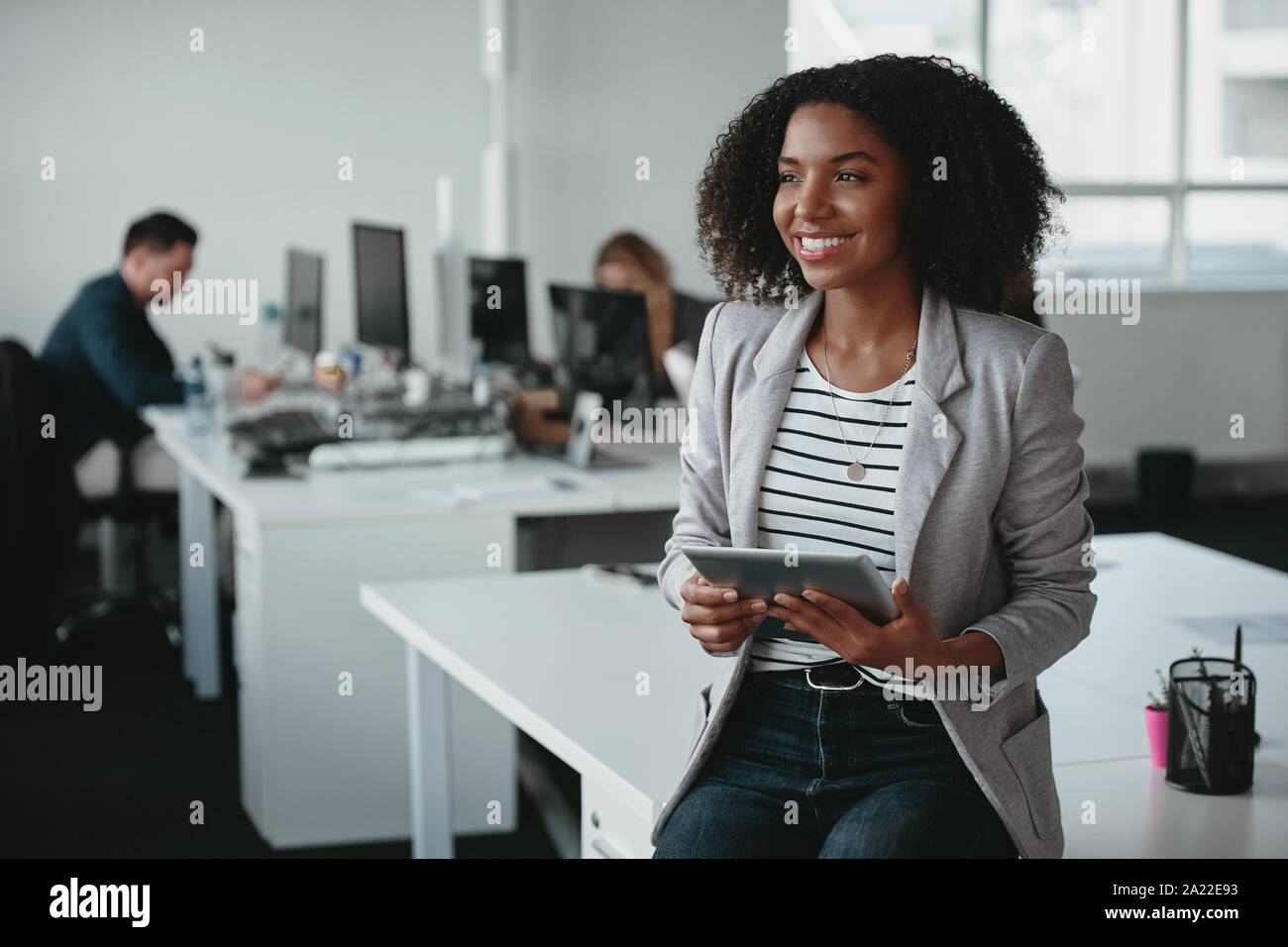 Felice pensieroso giovane imprenditrice con tavoletta digitale in mano e sorridente che guarda lontano nella parte anteriore del collega a sfondo Foto Stock