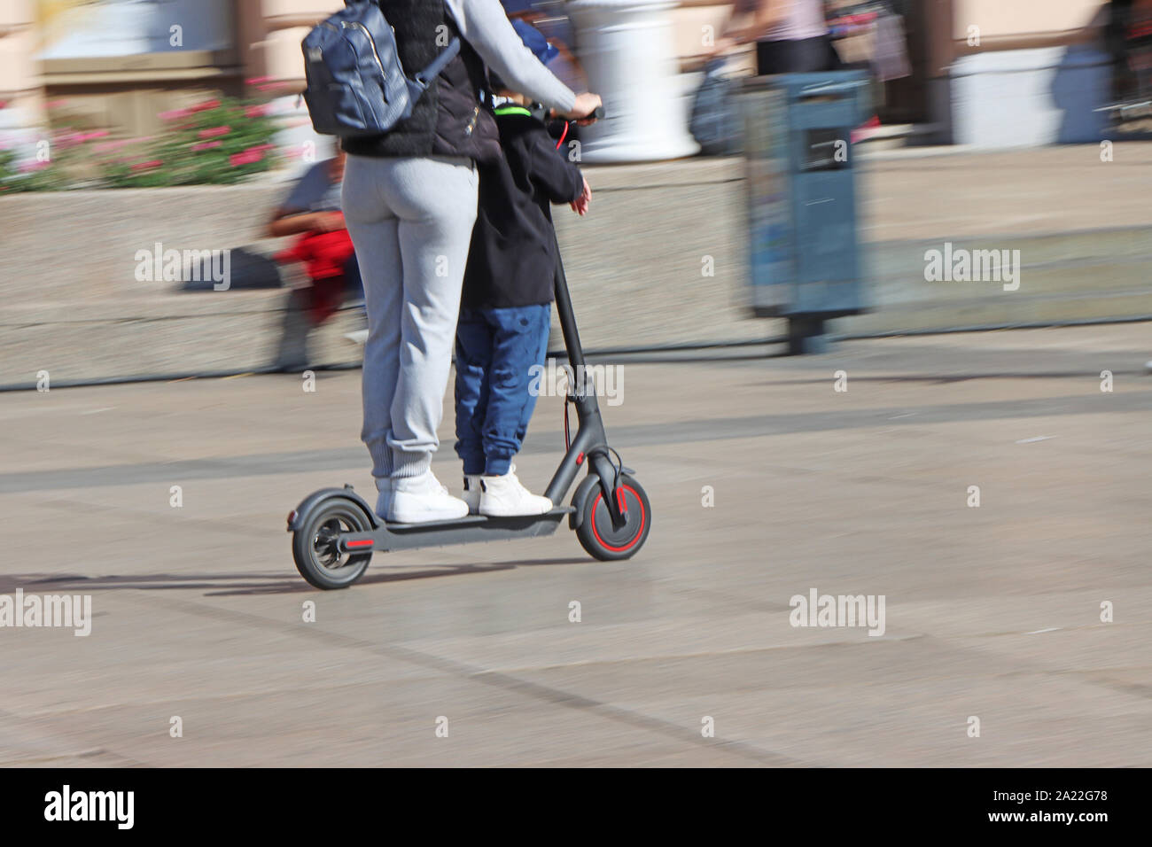 Madre e figlio di equitazione calcio elettrico scooter presso la piazza della città, movimento sfocato Foto Stock