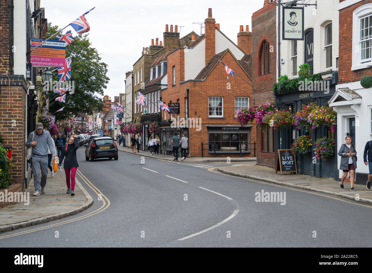 La gente fuori e circa in High Street, Eton. Scena colorata con fiore riempito nei cestini appesi e Union Jack Flag. Foto Stock