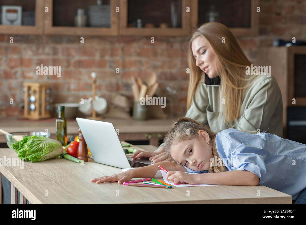 Figlia Blondy sensazione annoiato, madre parla al telefono e guardando il laptop, cucina interno, spazio di copia Foto Stock