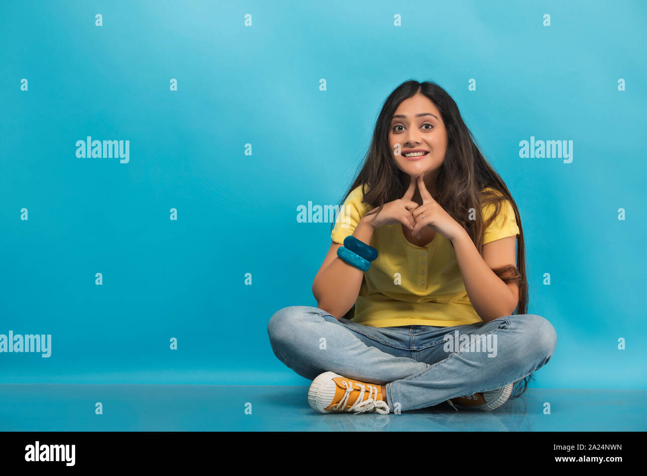 Sorridente ragazza adolescente in jeans e maglietta seduta zampe trasversale sul pavimento con le sue mani toccano il suo mento Foto Stock