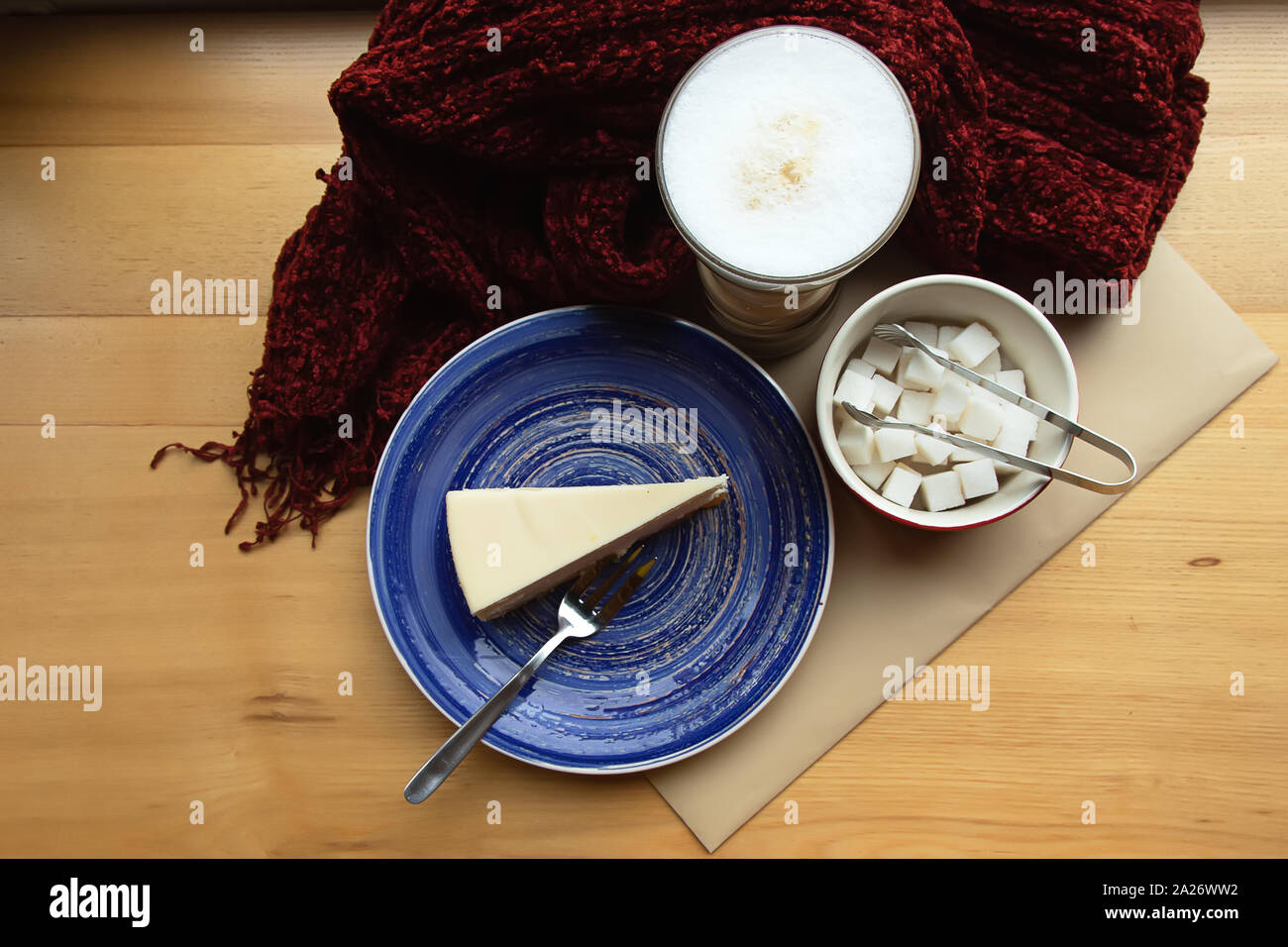 Un pezzo di torta di formaggio su una piastra di blu, forcella, zucchero, tazza di cappuccino e sciarpa rossa su un tavolo di legno Foto Stock