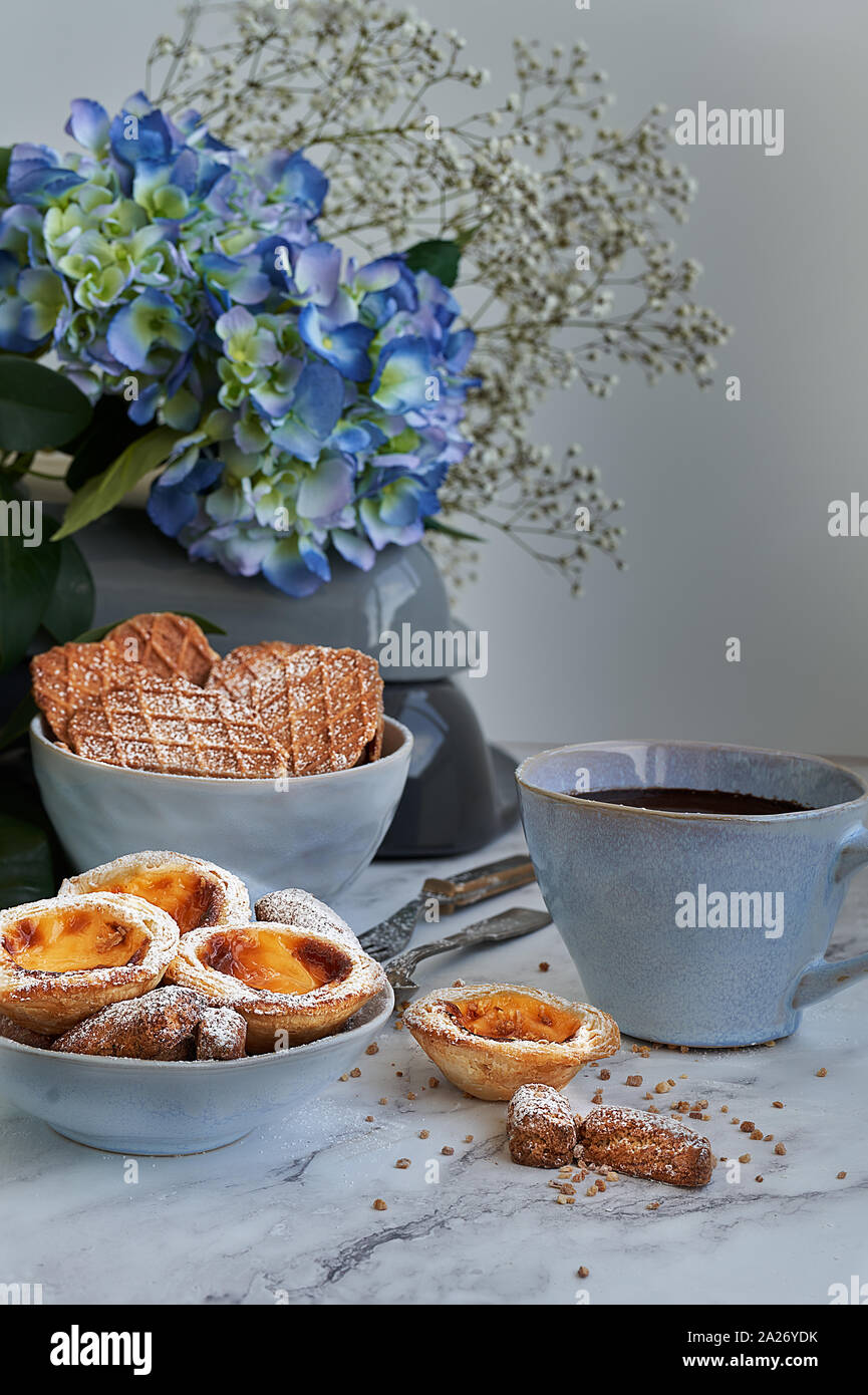 Pasta sfoglia e crema inglese dolci accompagnati da una tazza di cioccolato. Marmo bianco sullo sfondo blu e fiori. Foto Stock