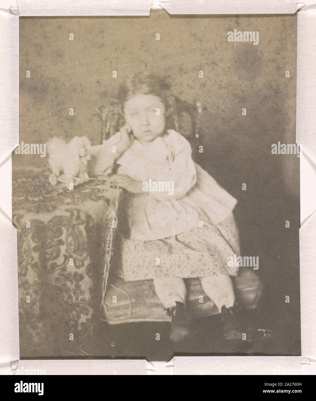 Ritratto di una ragazza non identificato, a piena lunghezza ritratto in studio, rivolto verso la parte anteriore, seduto con il gomito destro appoggiato su un tavolo e con la sua mano sinistra sul bordo del tavolo, accanto a un cane di pull-giocattolo su ruote Foto Stock