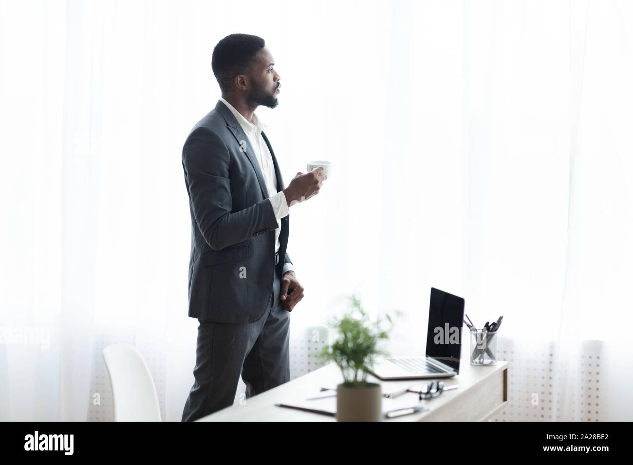 Malinconici imprenditore in piedi vicino alla finestra in ufficio e di bere il caffè Foto Stock