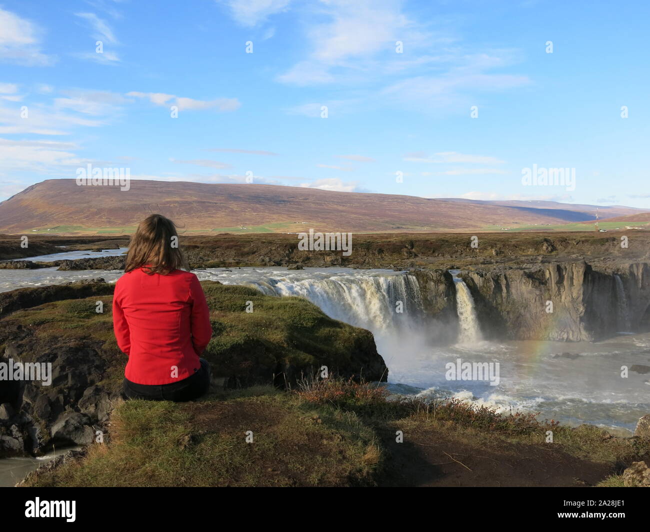 Una giovane donna si siede su una roccia e guarda fuori il vasto paesaggio vulcanico del Nord Islanda e le cascate Godafoss. Foto Stock