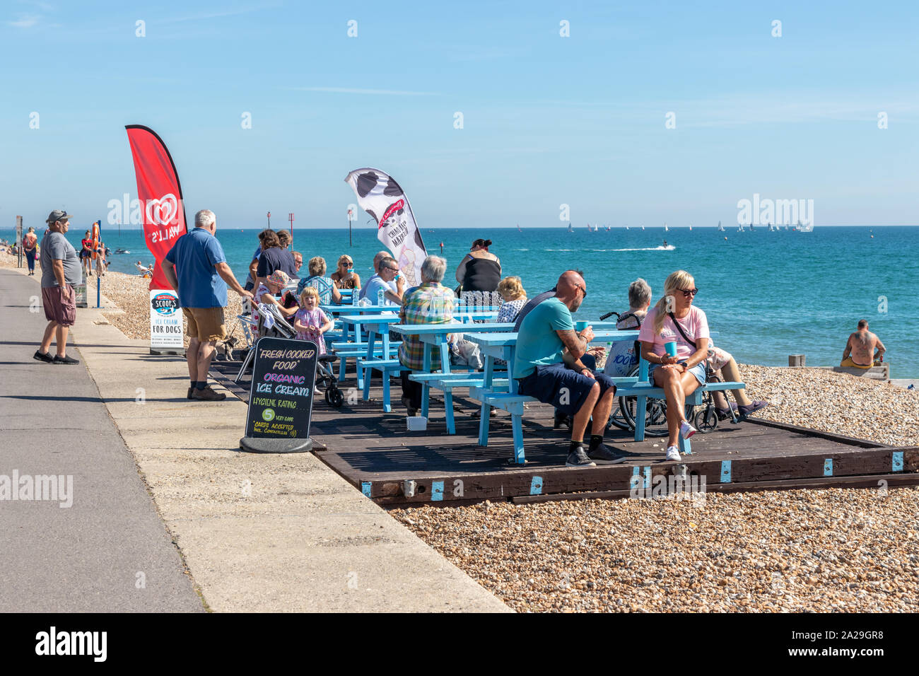 Turisti seduti su panchine fuori da un caffè sul lungomare gustando gelati e cibo in una calda giornata estiva A Bognor West Sussex Foto Stock