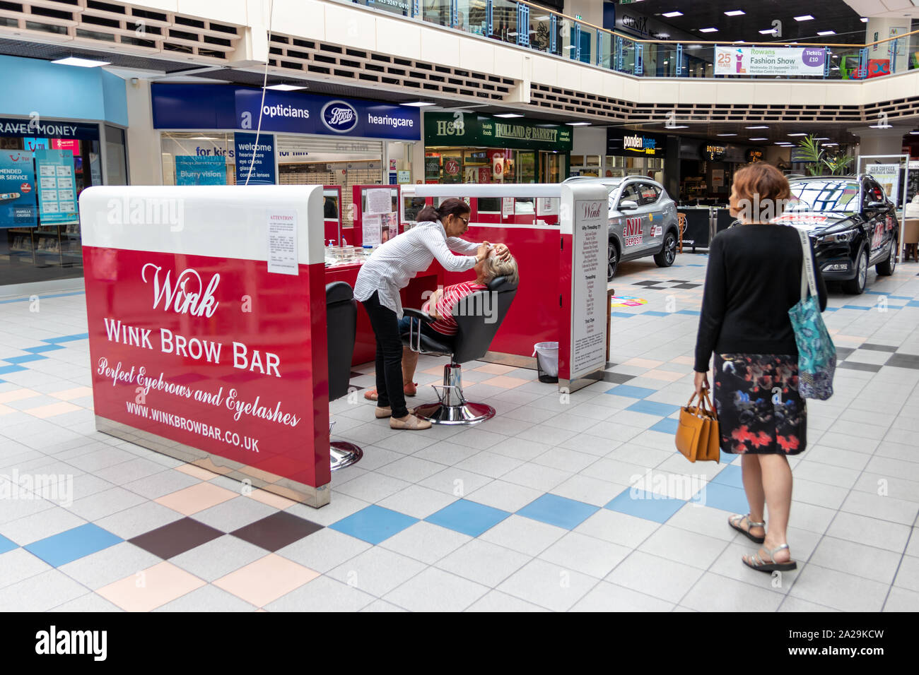 Una donna di mezza età a guardare come il suo amico ha le sopraciglia filettato in un centro commerciale o centro commerciale Foto Stock