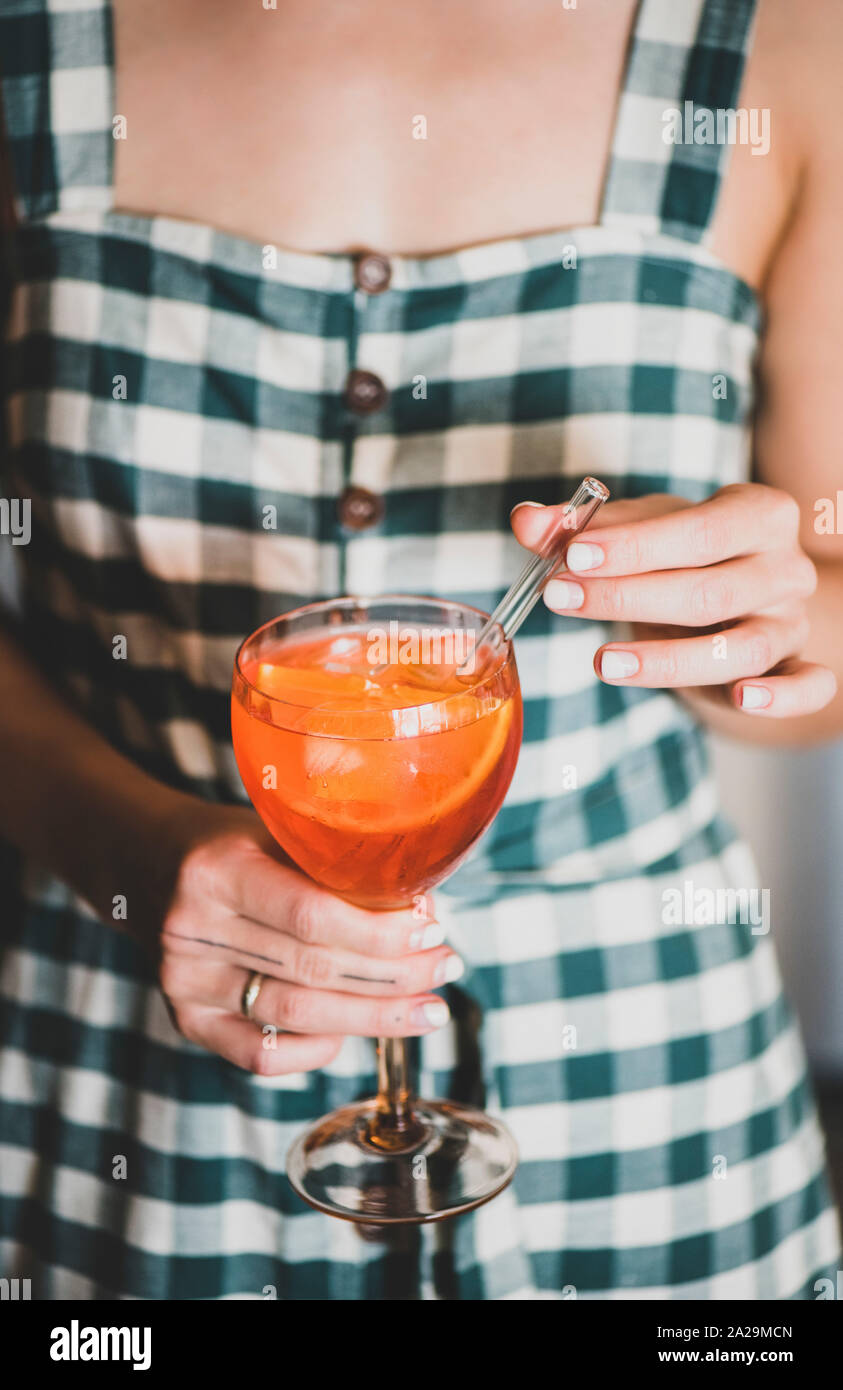 Giovane donna in abito a scacchi tenendo un bicchiere di Aperol Spritz cocktail drink con orange in vetro. Estate bevanda rinfrescante concetto Foto Stock