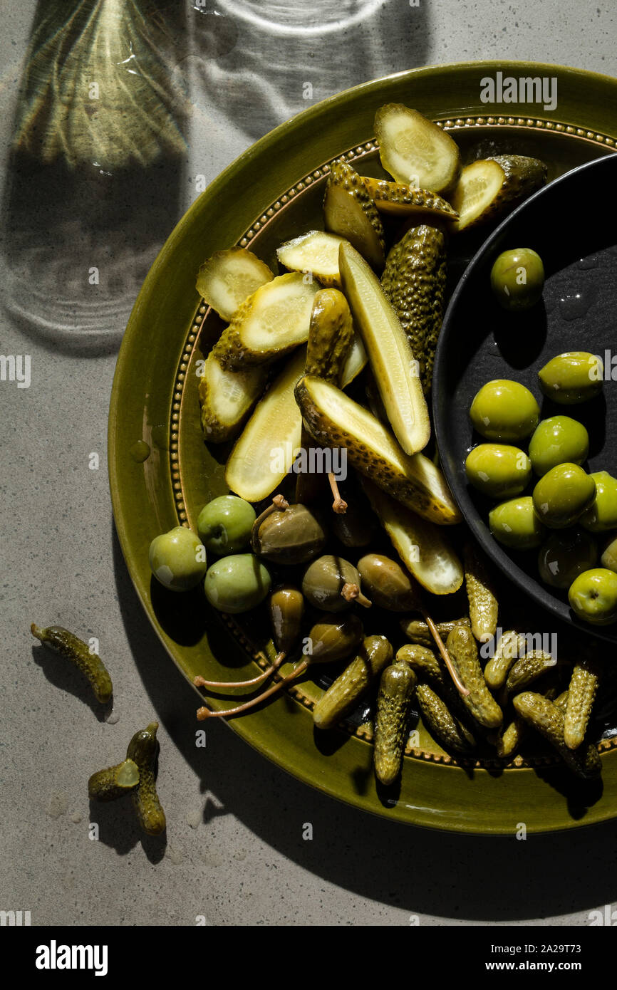 Una selezione di cetriolini sottaceto, bacche di capperi e olive verdi su un verde oliva piastra. Ombre sullo sfondo di calcestruzzo suggeriscono di bicchieri di vino. Foto Stock