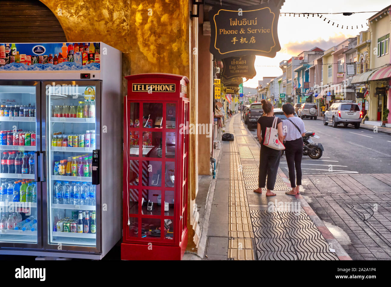 I turisti stand in Thalang Rd. nella città di Phuket, Thailandia, vicino ad un negozio che vende bevande fredde e telefoni cellulari mantenute in una simulazione di British Phone Booth Foto Stock