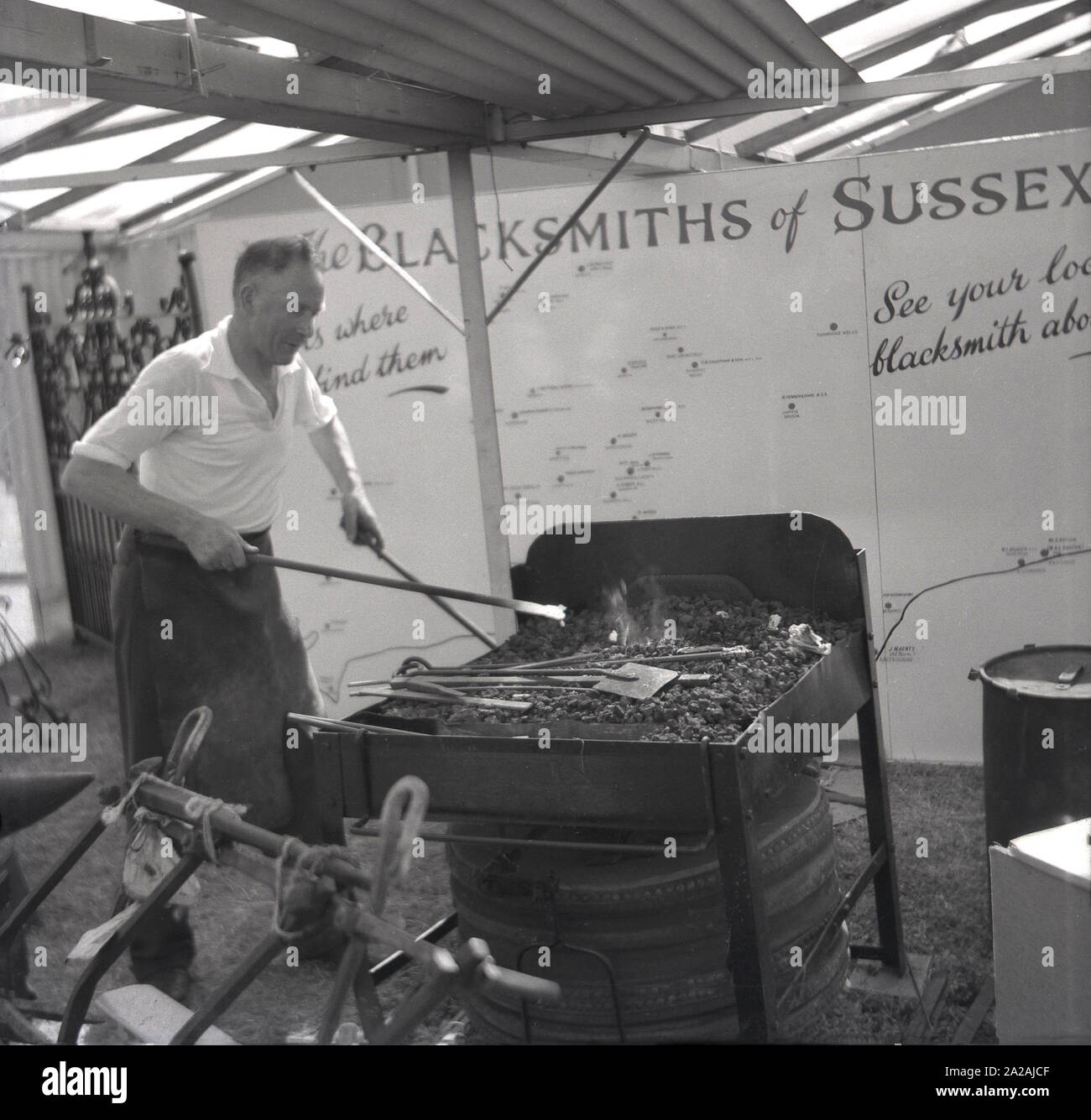 Anni '1950, storico, un fabbro che dimostrava le sue abilità di lavorazione del ferro su una forgia in una tenda in una mostra agricola, promuovendo le abilità dei contadini artigiani o dei metalmeccanici, Sussex, Inghilterra, Regno Unito. Nella foto si può vedere una grande tavola che mostra le posizioni dei diversi fabbri del Sussex. Foto Stock
