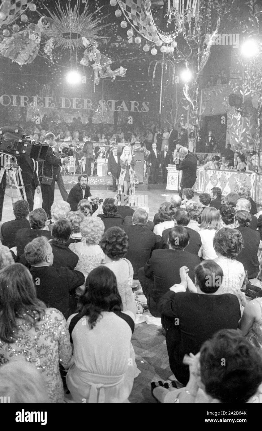 Vista della sala da ballo durante il programma. La foto mostra una donna sul palco e il pubblico da dietro. Dal soffitto pendono lampade e ornamenti. Nella terra di mezzo a sinistra è una telecamera. Sullo sfondo il lettering Soiree der Stelle. Foto Stock