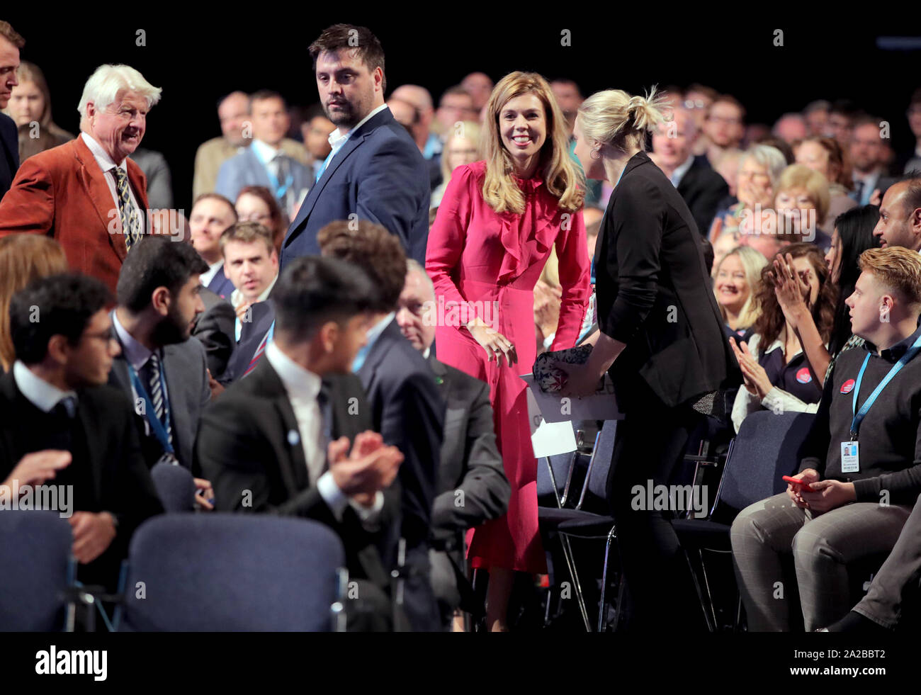 Carrie Symonds (al centro in rosso) seguita da Stanley Johnson (sinistra), il padre del Primo Ministro Boris Johnson, prendere i loro posti a sedere davanti al suo intervento durante il congresso del partito conservatore al Manchester Convention Center. Foto Stock