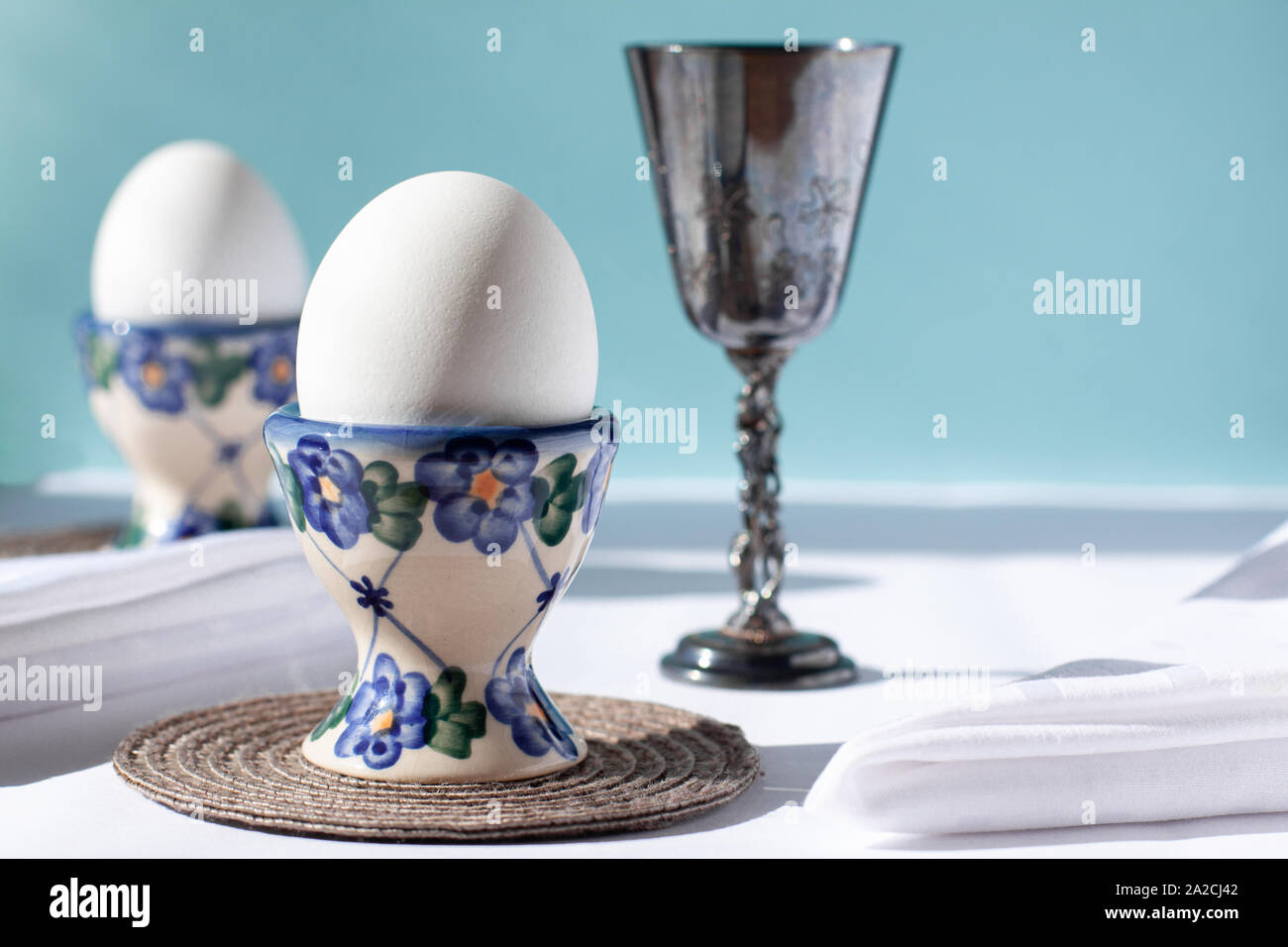 Ancora la vita di due uova bianche in gabbie di uovo con tovaglioli bianco vicino ad esso e Sterling Silver bicchiere di vino al giorno la luce del sole. Mondo internazionale delle uova al giorno Foto Stock