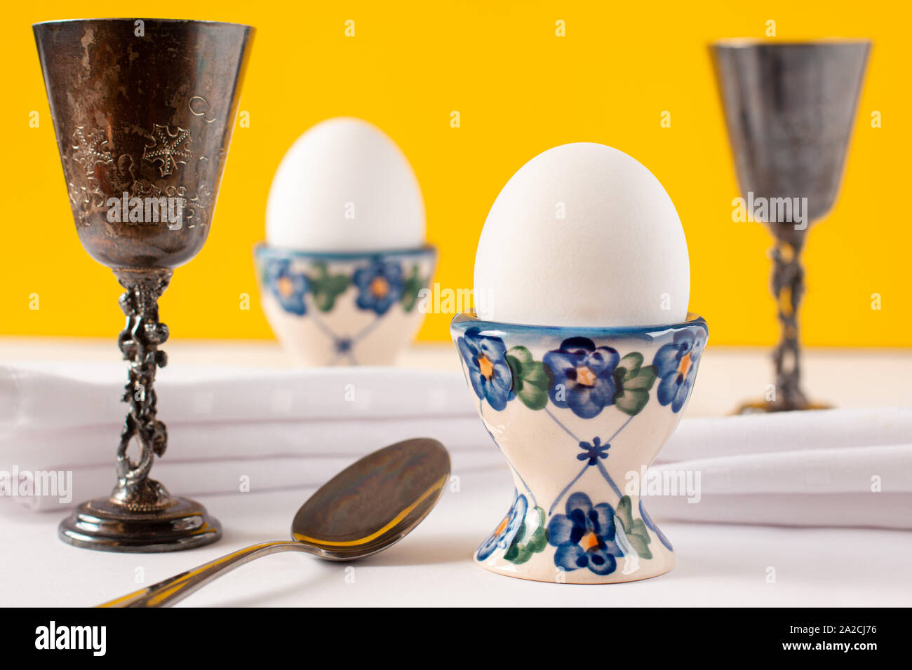 Ancora la vita di due uova bianche in gabbie di uovo con cucchiaio, tovaglioli bianco vicino ad esso e due argento sterling bicchieri di vino al giorno la luce del sole Foto Stock