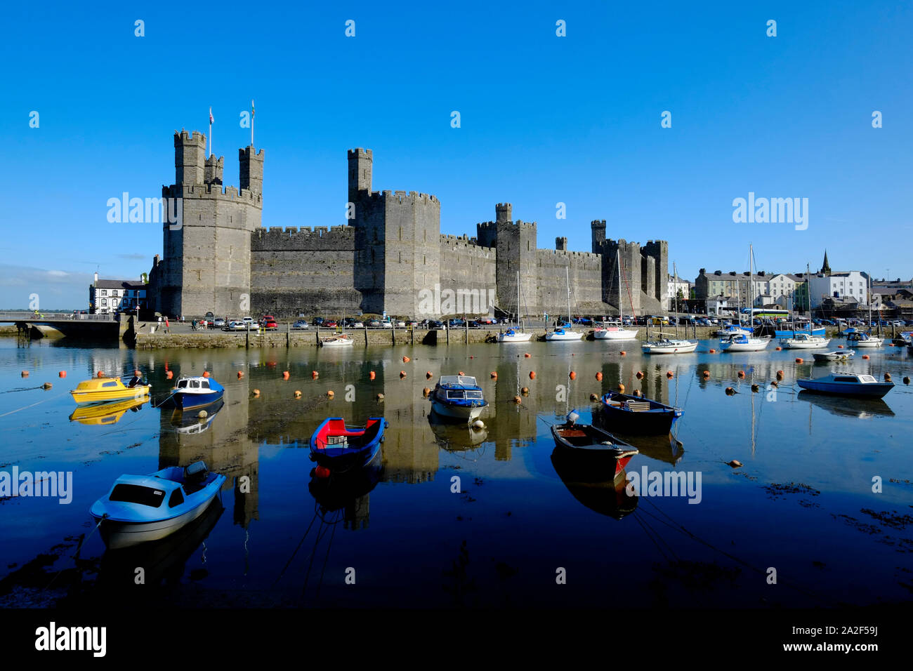 Vista sul Fiume Sieont verso Caernarfon Castle su ancora un calmo mattino riflettendo sull'acqua ad alta marea con barche ormeggiate in primo piano Foto Stock