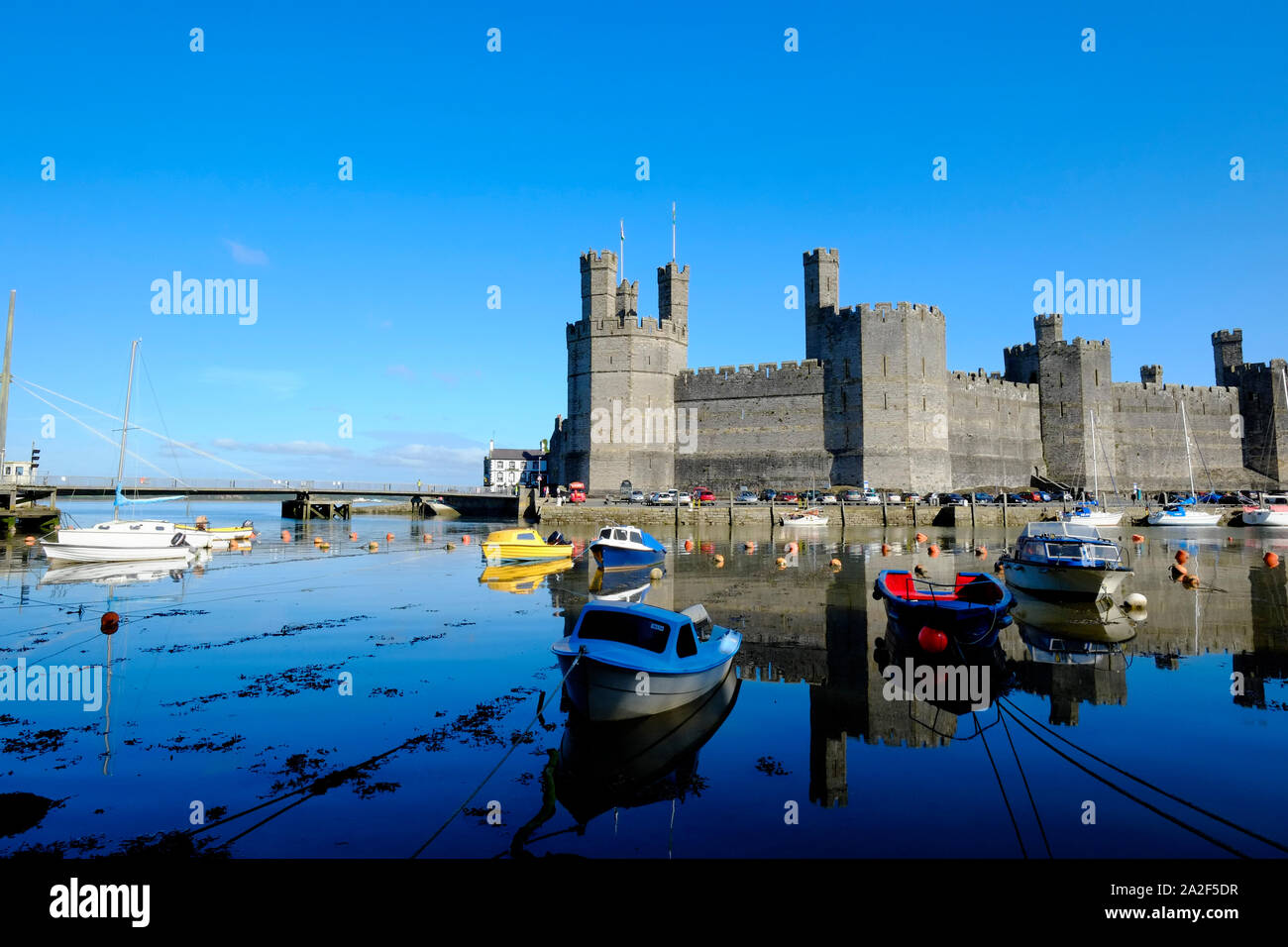 Vista sul Fiume Sieont verso Caernarfon Castle su ancora un calmo mattino riflettendo sull'acqua ad alta marea con barche ormeggiate in primo piano Foto Stock