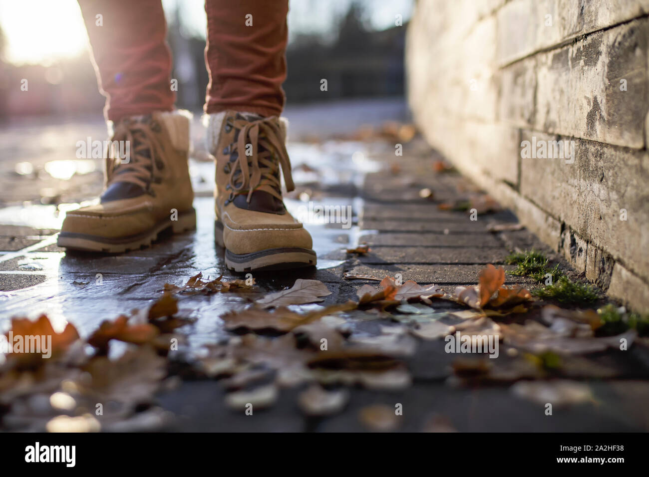 Gambe in stivali di stand su una pavimentazione in ciottoli di pietra in una pozza ricoperta di ghiaccio sotto i raggi del sole, intorno sono caduto foglie. Close-up. Foto Stock