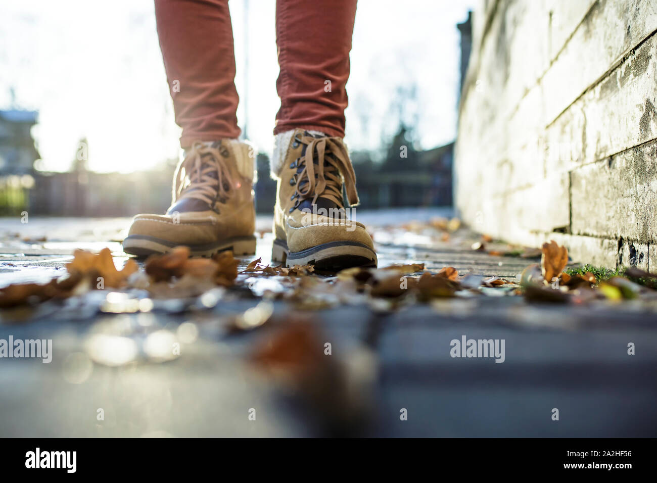 Gambe in stivali di stand su una pavimentazione in ciottoli di pietra in una pozza ricoperta di ghiaccio sotto i raggi del sole, intorno sono caduto foglie. Close-up. Foto Stock