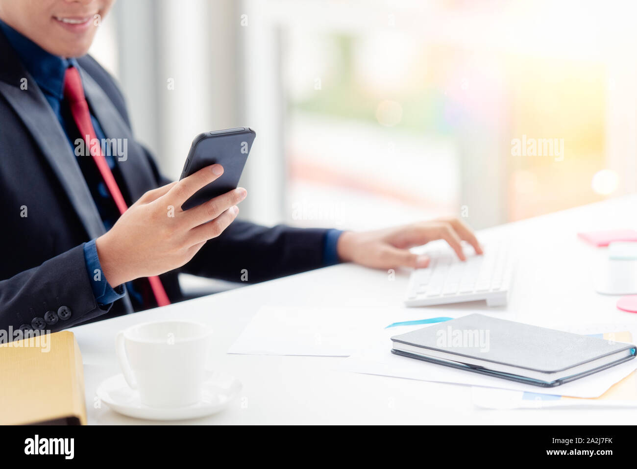 Un giovane imprenditore con un fiducioso suit seduti ad una scrivania in un ufficio moderno. Egli è in possesso di un telefono da contattare per il lavoro mentre utilizzando un computer. Foto Stock