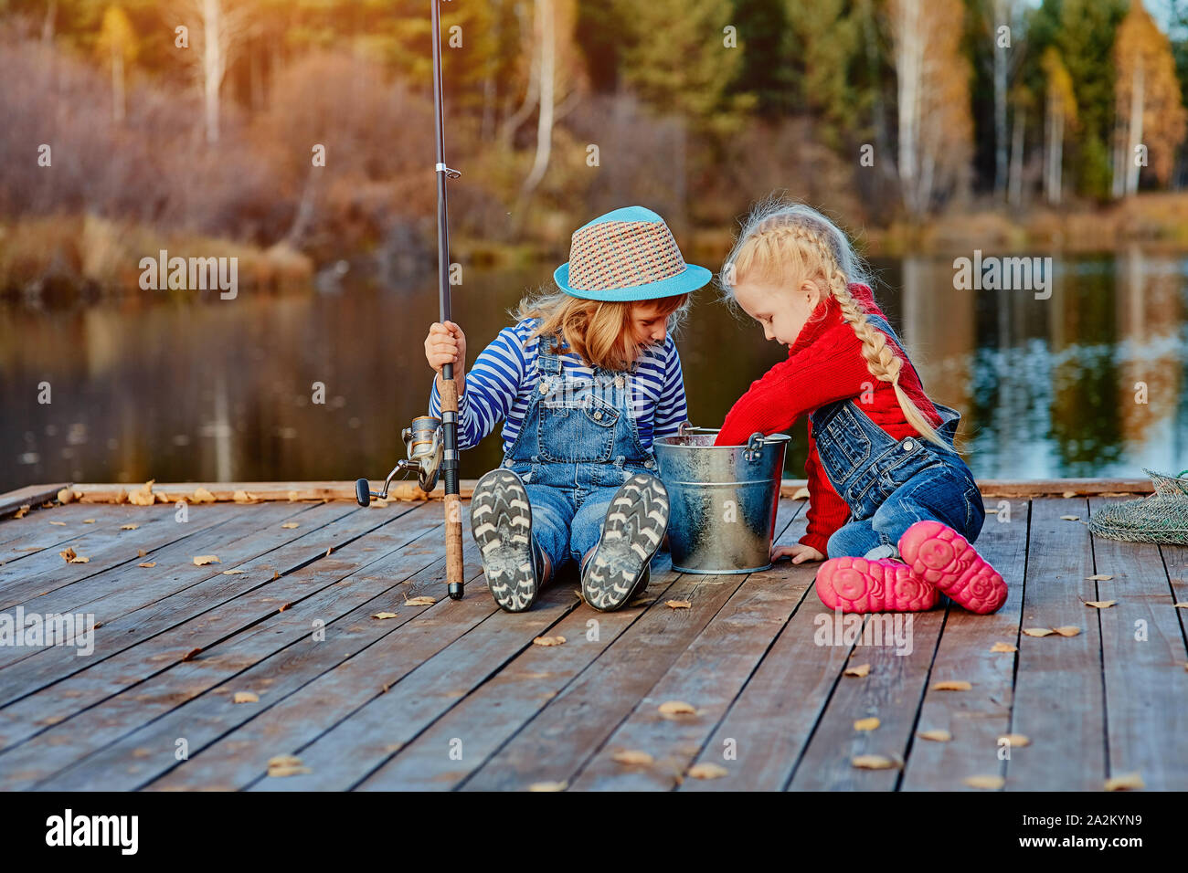 Due piccole sorelle o amici siedono con canne da pesca su un molo in legno. Hanno catturato un pesce e metterlo in un secchio. Sono felici con la loro cattura e d Foto Stock