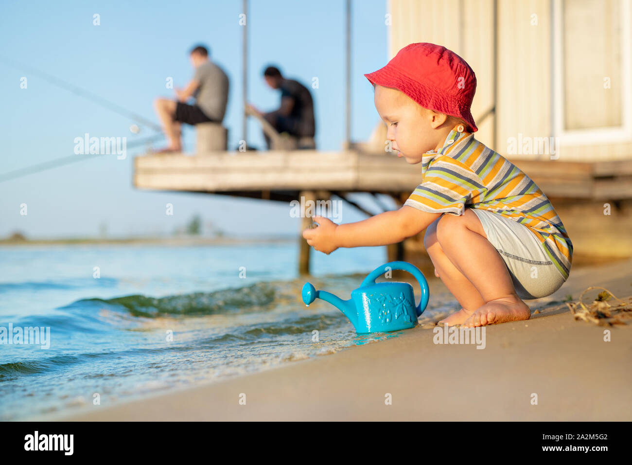 Carino adorabile bionda caucasica toddler boy divertendosi con blue annaffiatoio al fiume o lago di riva sabbiosa mentre il padre la pesca sullo sfondo Foto Stock