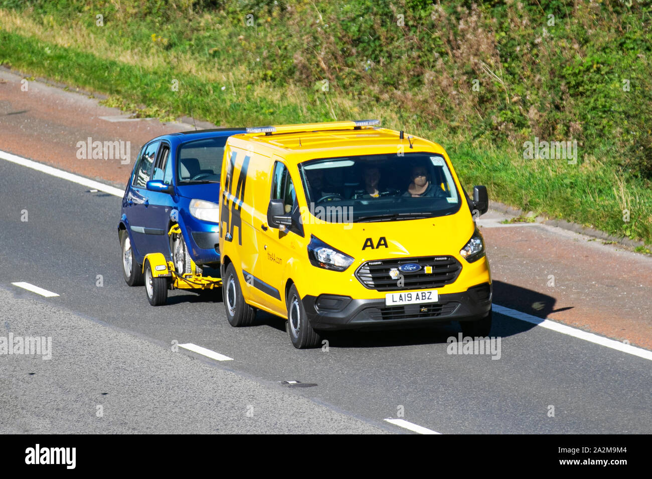 Pulmino giallo AA per il recupero di 24 ore, Ford Transit Custom 340 per il trasporto di base del veicolo guasto. Vista laterale del veicolo di recupero guasti che trasporta una piccola auto per famiglie blu lungo la M6, Lancaster, Regno Unito; traffico veicolare, trasporto, moderno, in direzione nord sull'autostrada a 3 corsie. Foto Stock