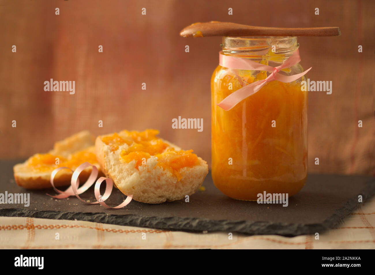 Vaso in casa di marmellata di arance con un pezzo di pane con la marmellata e uno sfondo marrone per far corrispondere la marmellata e lo spazio per i testi. Spazio di copia Foto Stock