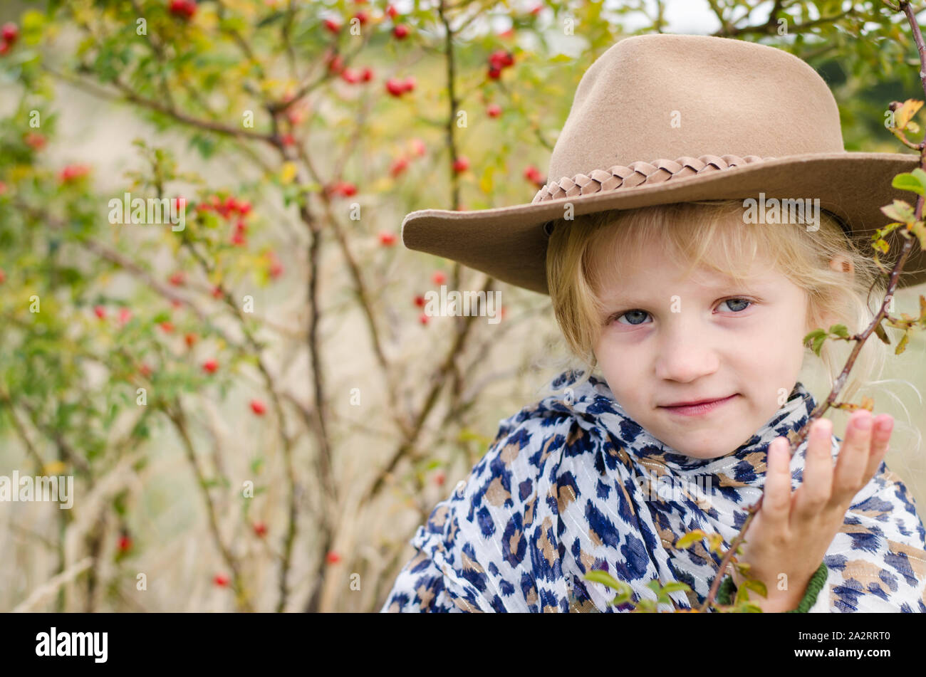 Bella sorridente ragazza bionda con marrone cappello nella sua testa in autunno campo con rose-hip impianto Foto Stock